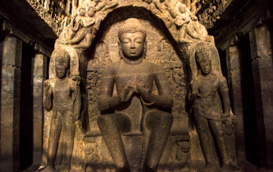A stone Buddha statue in a temple with intricate carvings and two standing figures on either side.