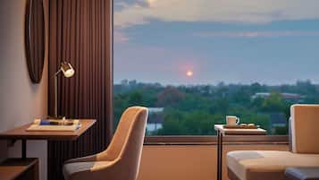 Una habitación de hotel moderna con un escritorio, una silla y una pequeña mesa con una taza y galletas. Un ventanal grande ofrece vistas de un atardecer sobre un paisaje urbano.
