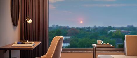 Una habitación de hotel moderna con un escritorio, una silla y una pequeña mesa con una taza y galletas. Un ventanal grande ofrece vistas de un atardecer sobre un paisaje urbano.