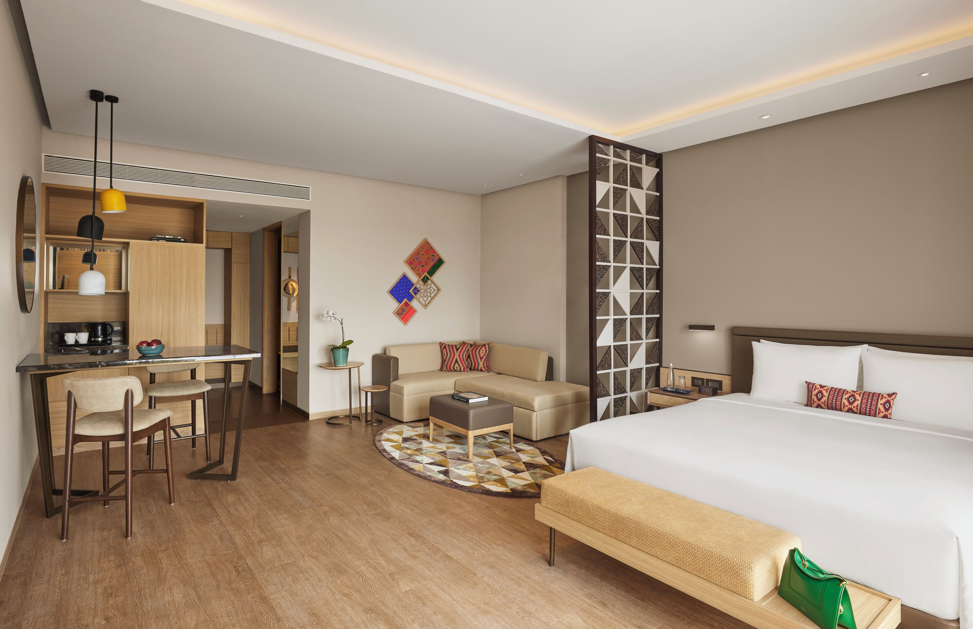 A hotel room with a bed, a sofa, a desk with chairs, a coffee machine, and a small table with a plant.