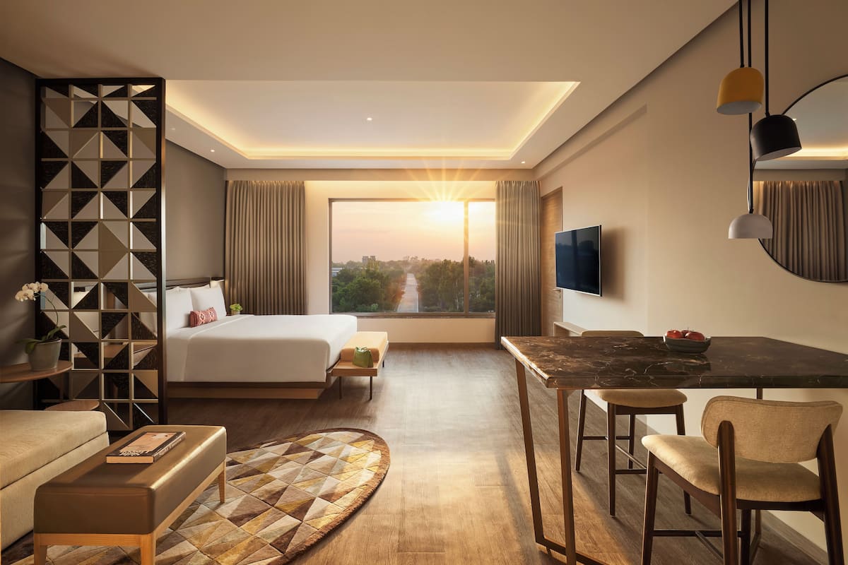 Ein modernes Hotelzimmer mit einem großen Bett, einem Schreibtisch mit Stühlen, einem Fernseher und Blick auf die Skyline im Abendrot.