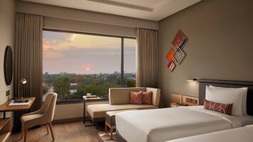 A hotel room with a large window, a desk, a sofa, and two beds.