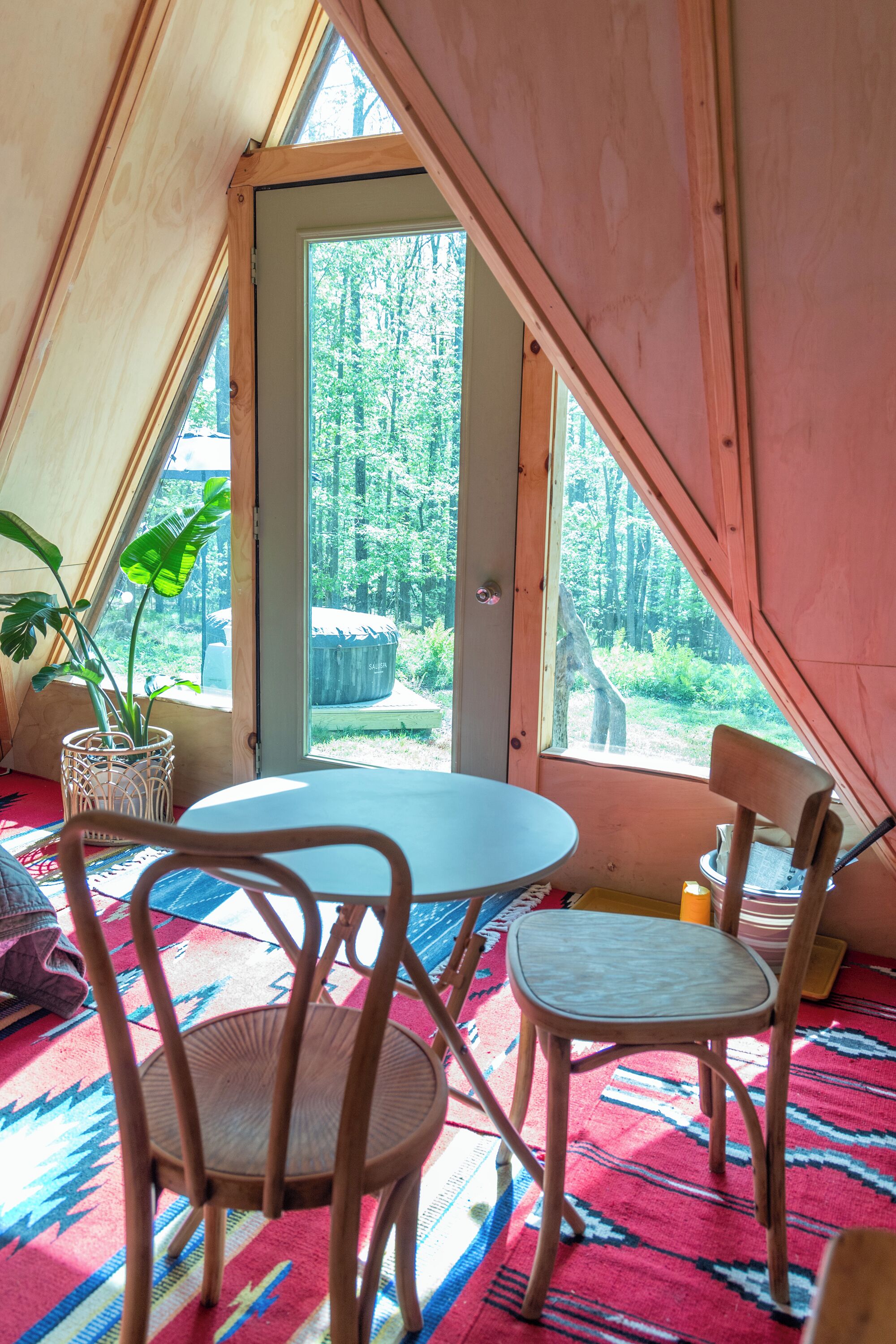 Una habitación acogedora con una mesa redonda, dos sillas de madera, una planta en maceta y vistas a un bosque a través de un ventanal grande.