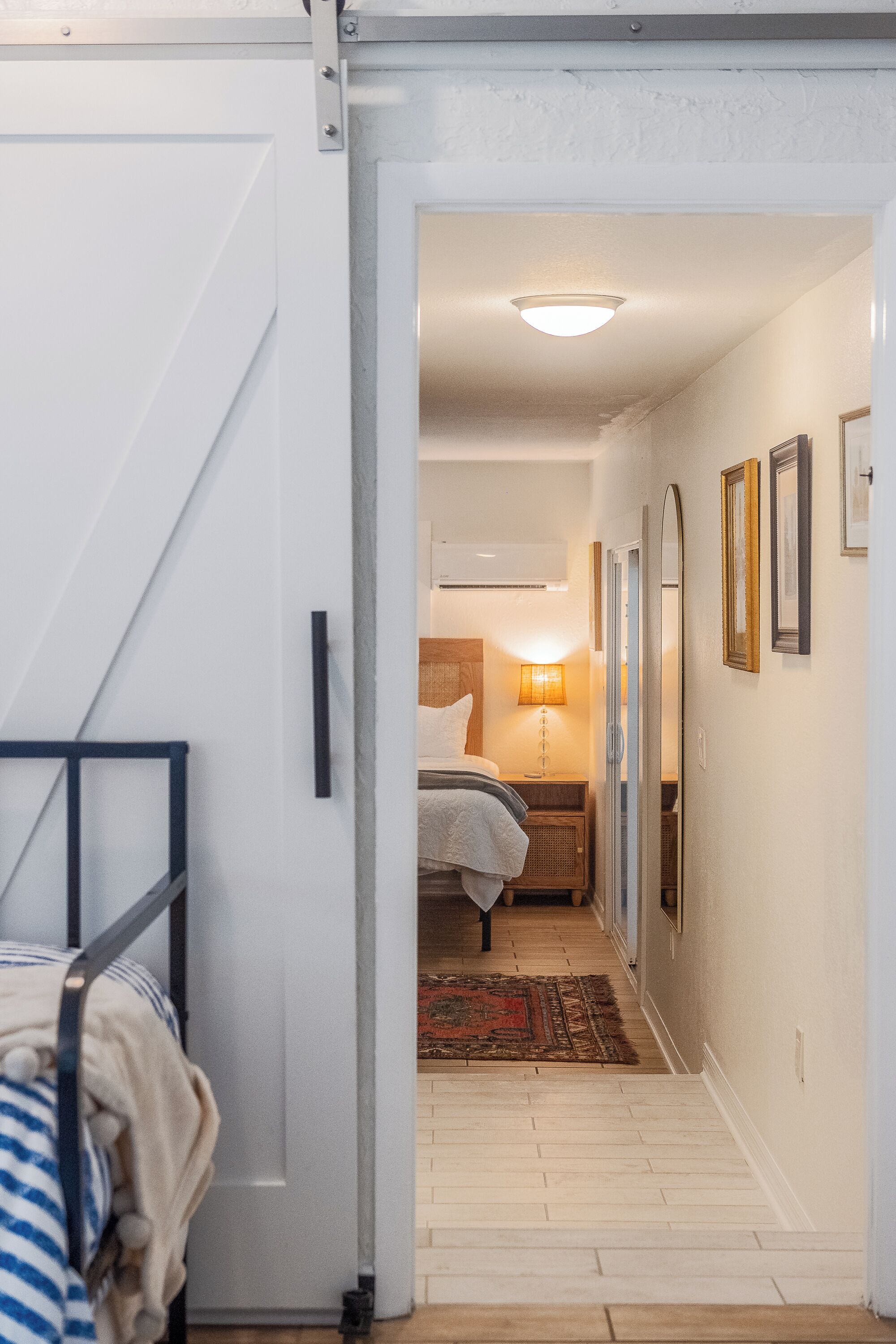 Ein Flur, der in ein Schlafzimmer mit Bett, Nachttisch, Lampe und gerahmten Bildern an der Wand führt.