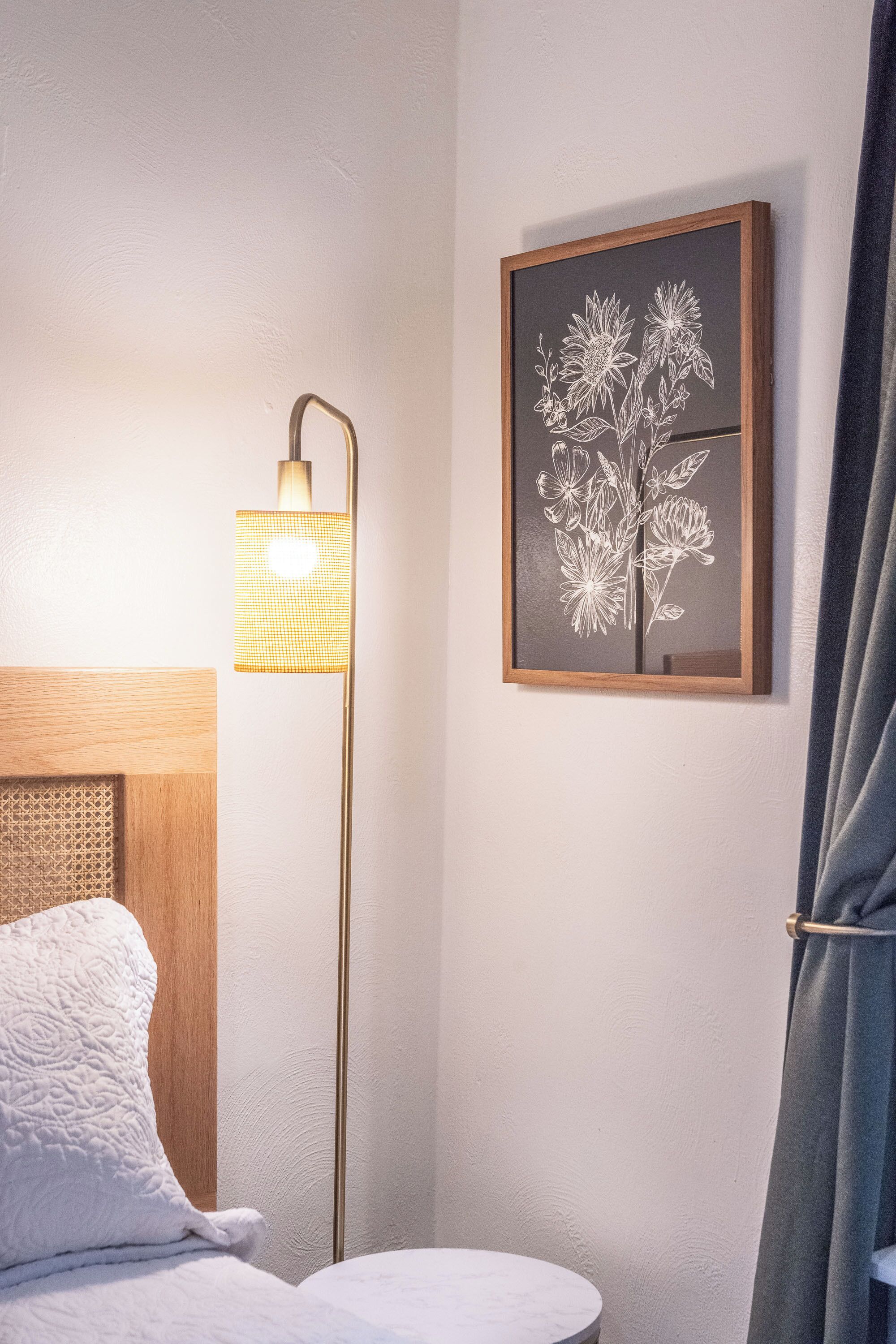 A bedroom with a bed, a bedside lamp, a framed floral artwork, and a curtain.