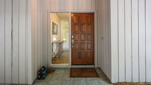 Uma porta de madeira com um painel de vidro, um tapete na entrada e uma pequena mesa com uma cadeira em frente a um prédio com revestimento branco.