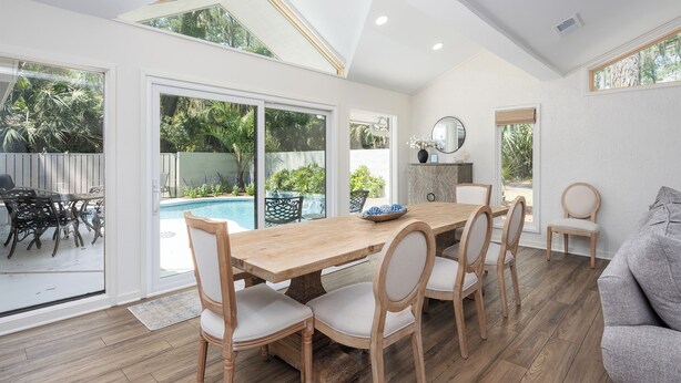 Uma sala de jantar com mesa e cadeiras de madeira, janelas amplas e vista para uma piscina e vegetação.