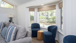 Quarto com um sofá cinzento, duas poltronas azuis, uma mesa lateral redonda de madeira e uma janela com portadas brancas.