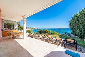 A covered outdoor area with a dining table and chairs, a poolside lounge area with sun loungers, and a view of the sea.