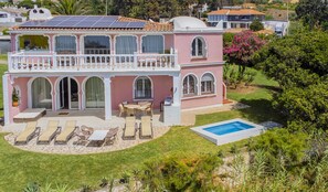 Una casa de dos pisos con balcón, piscina y patio con sillones de descanso.