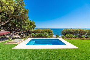 A rectangular swimming pool with a clear blue water surface, surrounded by well-maintained green grass, with a view of the sea and a tree-lined background.