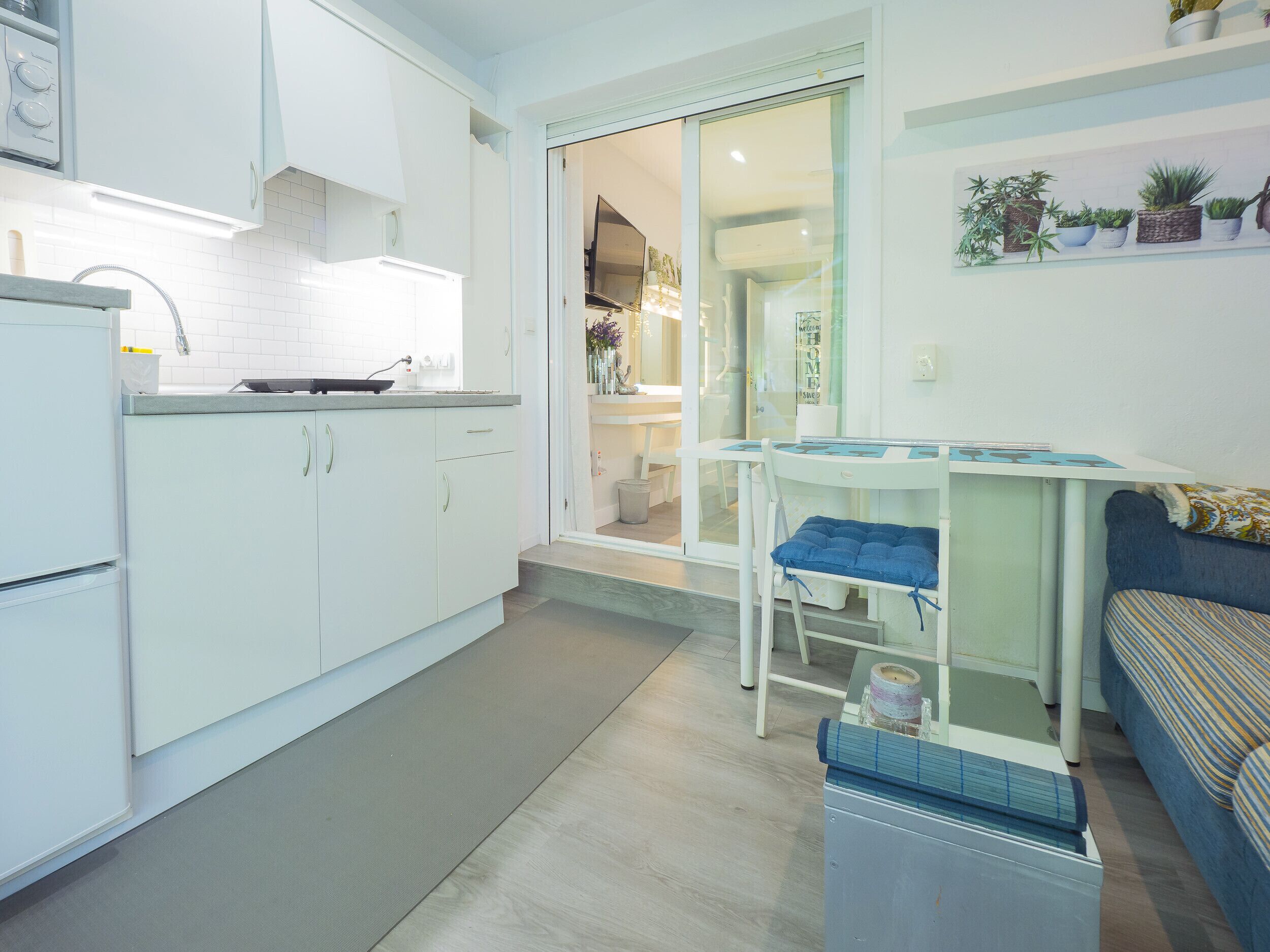 A compact kitchen with white cabinets, a sink, and a stove. There is a small table with a blue cushion and a shelf with potted plants.