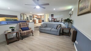 Un salon avec un canapé, un fauteuil et une petite table sur laquelle est posée une plante. On aperçoit un ventilateur de plafond et une cuisine au fond.