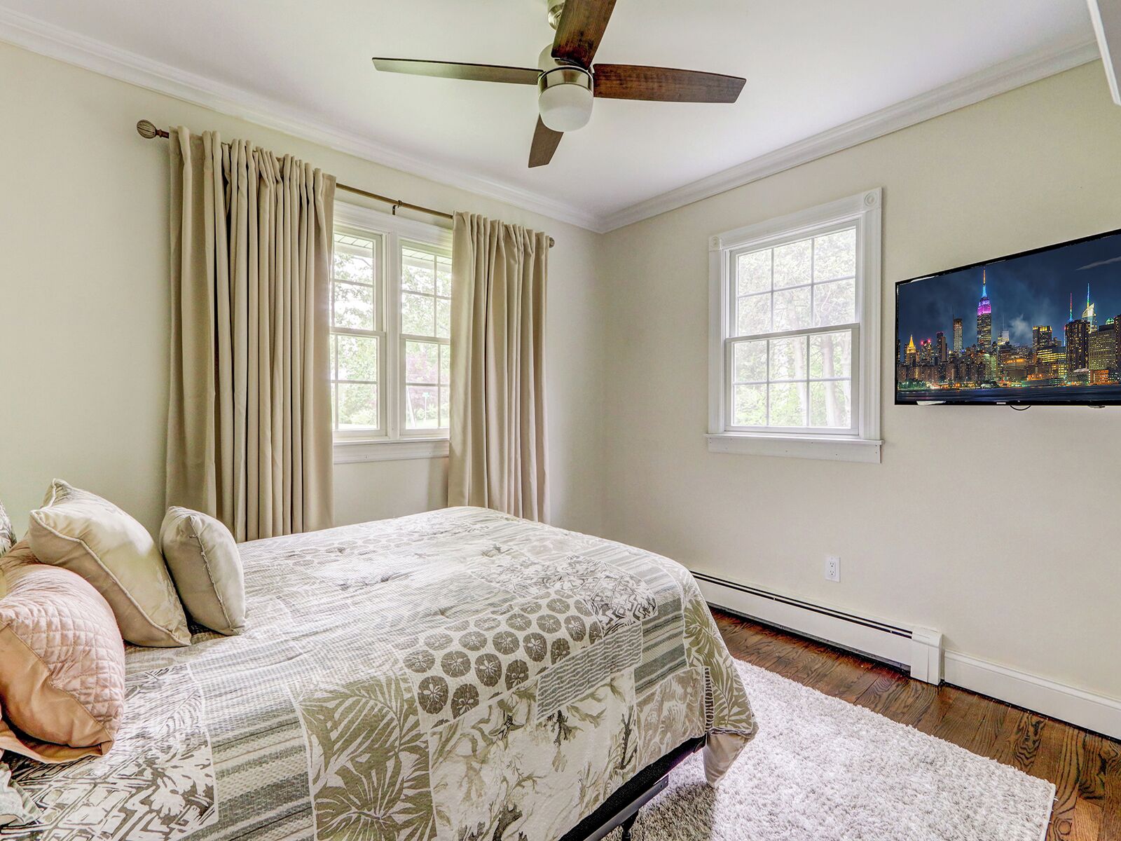 A bedroom with a bed, two windows, a ceiling fan, and a cityscape TV on the wall.