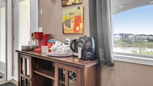 Camera d'albergo con una scrivania in legno, una macchina per il caffè, una ciotola di frutta e un angolo con bevande Tim Hortons. Due quadri alle pareti e vista su case dalla finestra.