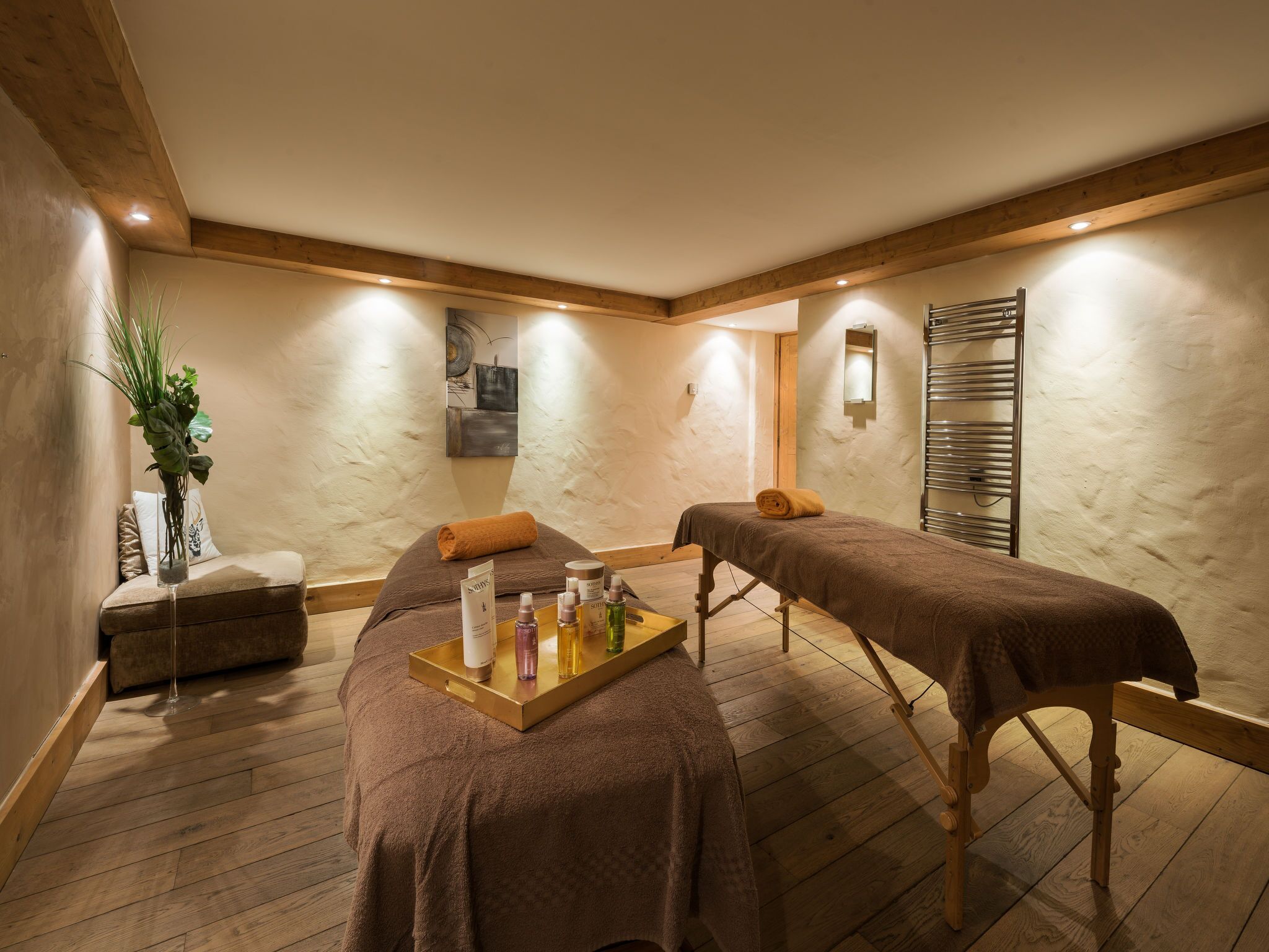 A spa room with two massage tables, a wooden bench, a tray with bottles, and a plant.