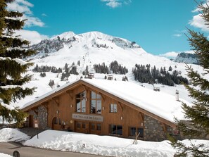 A wooden lodge with a stone facade, named "Le Village de Jessy," surrounded by snow-covered mountains and pine trees.