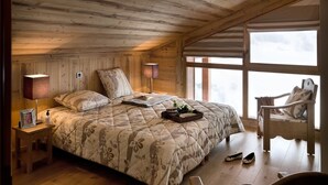 Une chambre confortable avec un plafond en bois, un grand lit recouvert d’une literie à motifs floraux, des lampes de chevet et une vue sur un paysage enneigé par la fenêtre.