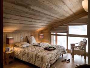 Een gezellige slaapkamer met een houten plafond, een groot bed met bloemenbedrukking, nachtlampjes en uitzicht op een besneeerd landschap door een raam.