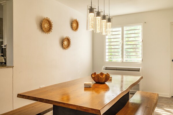 A wooden dining table with a bowl centerpiece, pendant lights, and wall decor in a bright room with a window.