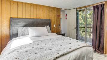 Une chambre à coucher avec une tête de lit en bois, un grand lit recouvert d’une courtepointe à motifs, une table de chevet et une porte donnant sur un balcon avec vue sur les arbres.