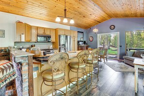A spacious kitchen with wooden cabinets, a large island with bar stools, and a view of the outdoors through a sliding glass door. - Woodsy Cabin Retreat ~ 4 Mi to Crescent Lake! (Crescent)