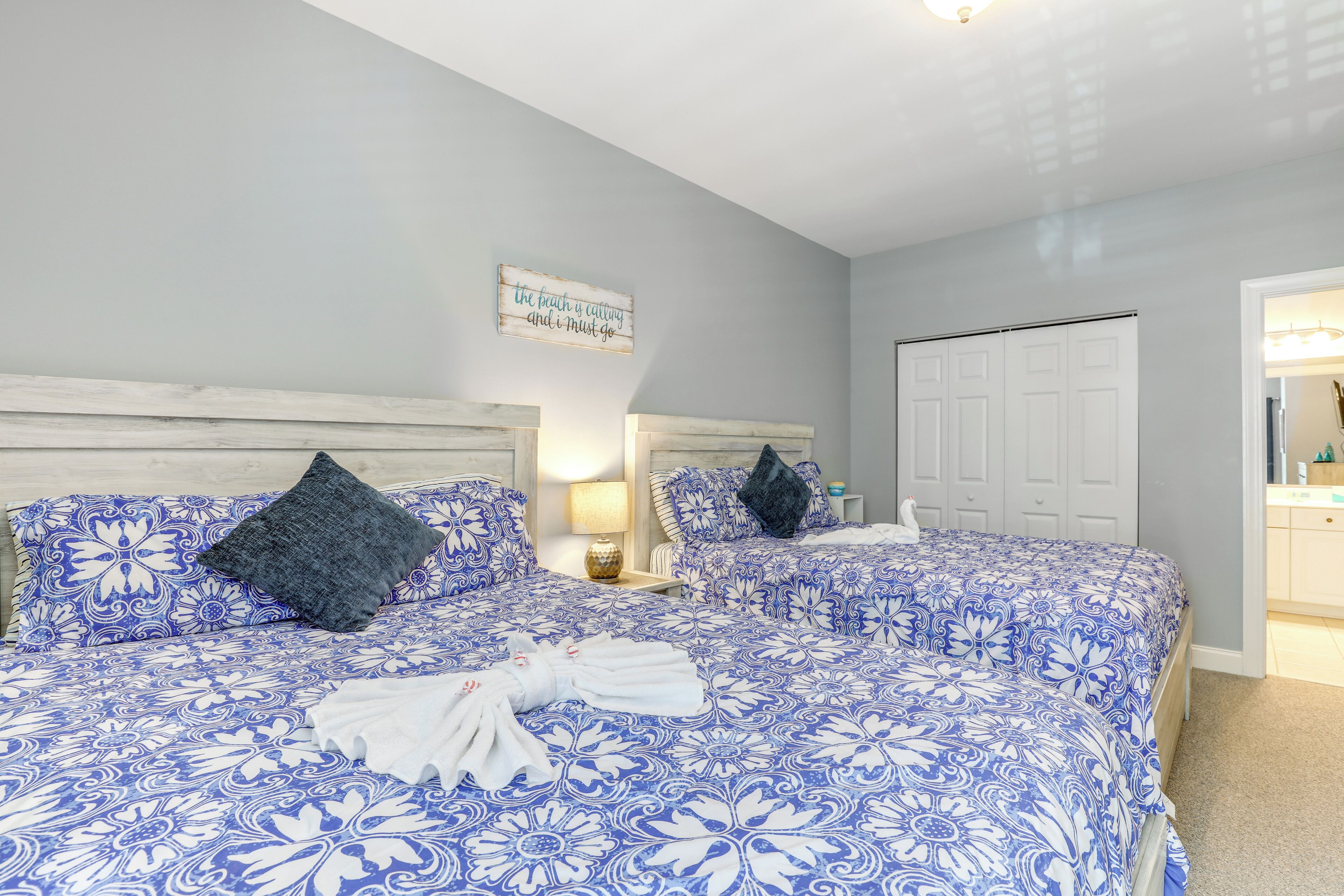 A bedroom with two beds, each with a blue floral bedspread and matching pillows. There is a wooden headboard, a nightstand with a lamp, and a wall-mounted sign.