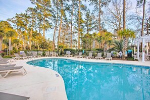 Una piscina amb gandules i una glorieta, envoltada per arbres alts i un cel serè.