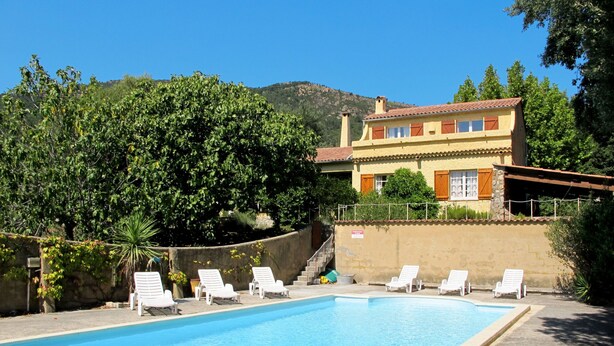 Une piscine entourée de chaises longues blanches, un bâtiment doté d’un toit en tuiles et un paysage montagneux en arrière-plan.