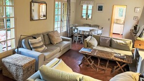 Un salon confortable avec un canapé, une petite table et une table à manger avec des chaises. Il y a des fenêtres avec des rideaux, un coin cuisine et une porte donnant sur l’extérieur.