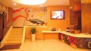 Un vestíbulo de hotel con mostrador de recepción, televisor, escalera y un gran mural.