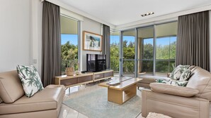 Ein modernes Wohnzimmer mit einer Couch, einem Couchtisch und einem Fernseher. Ein großes Fenster bietet Blick auf Bäume und einen Balkon.