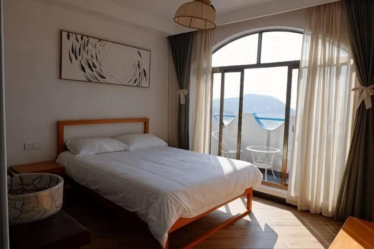 Ein Schlafzimmer mit Bett, Nachttisch, Lampe, einem Fenster mit Vorhängen und Meerblick.