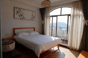 A bedroom with a bed, a bedside table, a lamp, a window with curtains, and a view of the sea. - Shensi Gouqi Island Mr.Tang's Private Garden Inn (Shengsi)