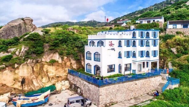 A multi-story building with a stone base and blue accents, situated on a hillside with a cliffside and a small dock area. - Shensi Gouqi Island Mr.Tang's Private Garden Inn (Shengsi)