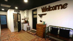 Una habitación con suelo de ladrillo, un banco de madera, un cartel de 'Welcome', un escritorio pequeño con una lámpara y un armario con la etiqueta 'Antiques'.