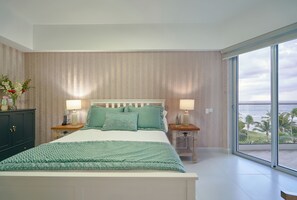 Quarto com cama grande, duas mesas de cabeceira com luminárias, cômoda e vista para o oceano através de grandes janelas.