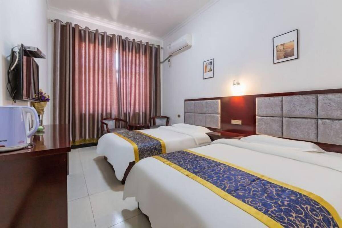 A hotel room with two beds, a desk with a kettle, a chair, a window with curtains, and a wall-mounted light.