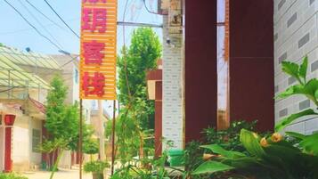 A narrow street with buildings on either side, a vertical sign with text, and some greenery in the foreground.