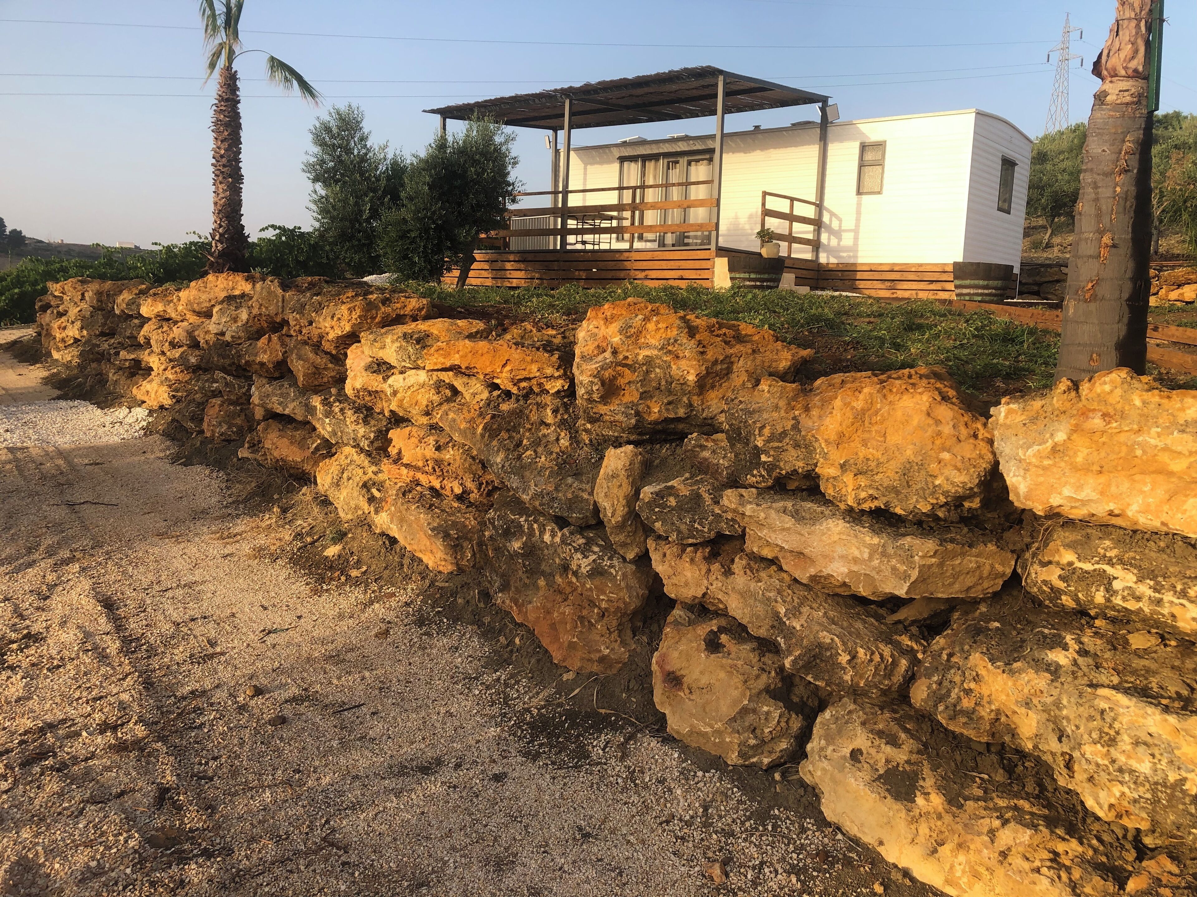 Un muro de piedra rústico, una casa móvil blanca con terraza de madera y una palmera.