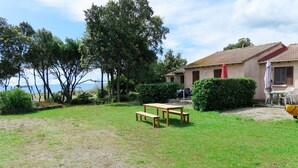 Un espace extérieur verdoyant avec une table et des bancs en bois, un bâtiment doté d’un toit en tuiles et une vue sur la mer.