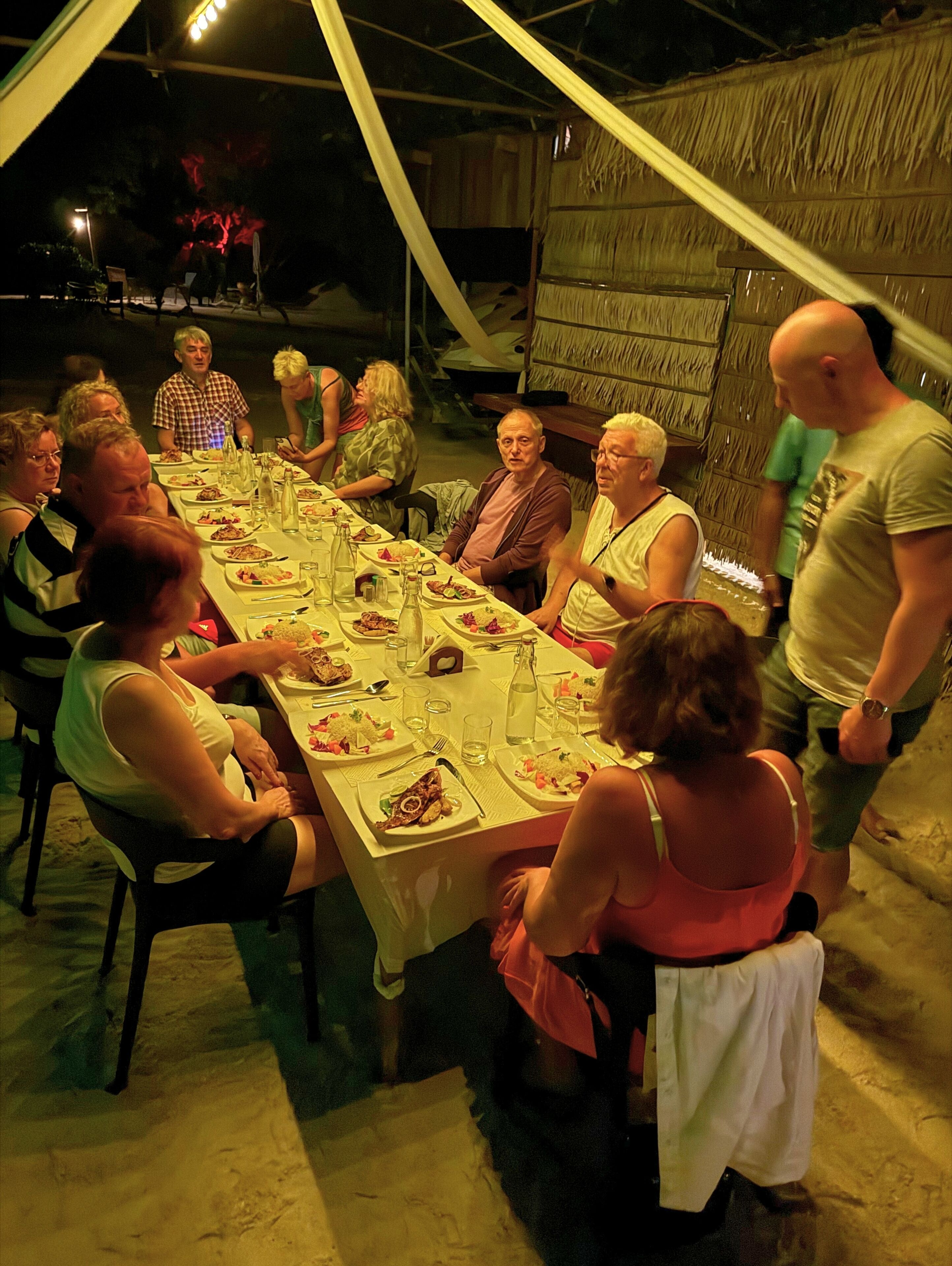 Een groep mensen zit rond een lange tafel met eten en drinken, onder een tentachtige constructie.