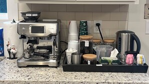 Una cocina con cafetera, tazas blancas apiladas, tarros de granos de café, hervidor y máquina de café.