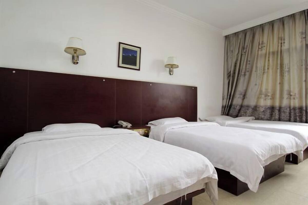 Hotellihuone, jossa on kolme erillistä sänkyä, puinen pääty, kaksi seinään kiinnitettyä lamppua, kehystetty taulu ja suuri ikkuna, jossa on verhot.