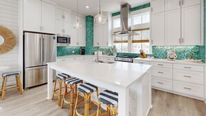 Une cuisine moderne avec des meubles blancs, un grand îlot central doté de tabourets, des appareils en acier inoxydable et un dosseret recouvert de carreaux verts.