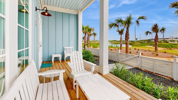 Un espace extérieur aménagé, abrité et doté de chaises Adirondack blanches et d’une table, offrant une vue sur un jardin paysager agrémenté de palmiers.
