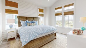 Une chambre à coucher avec un grand lit, une tête de lit en bois, des murs blancs et de nombreuses fenêtres équipées de stores.