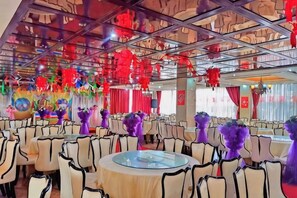 A banquet hall with round tables and chairs, red hanging decorations, and a colorful wall mural. - Zoige Grand Hotel (Zoige County People's Government) (Zoige)