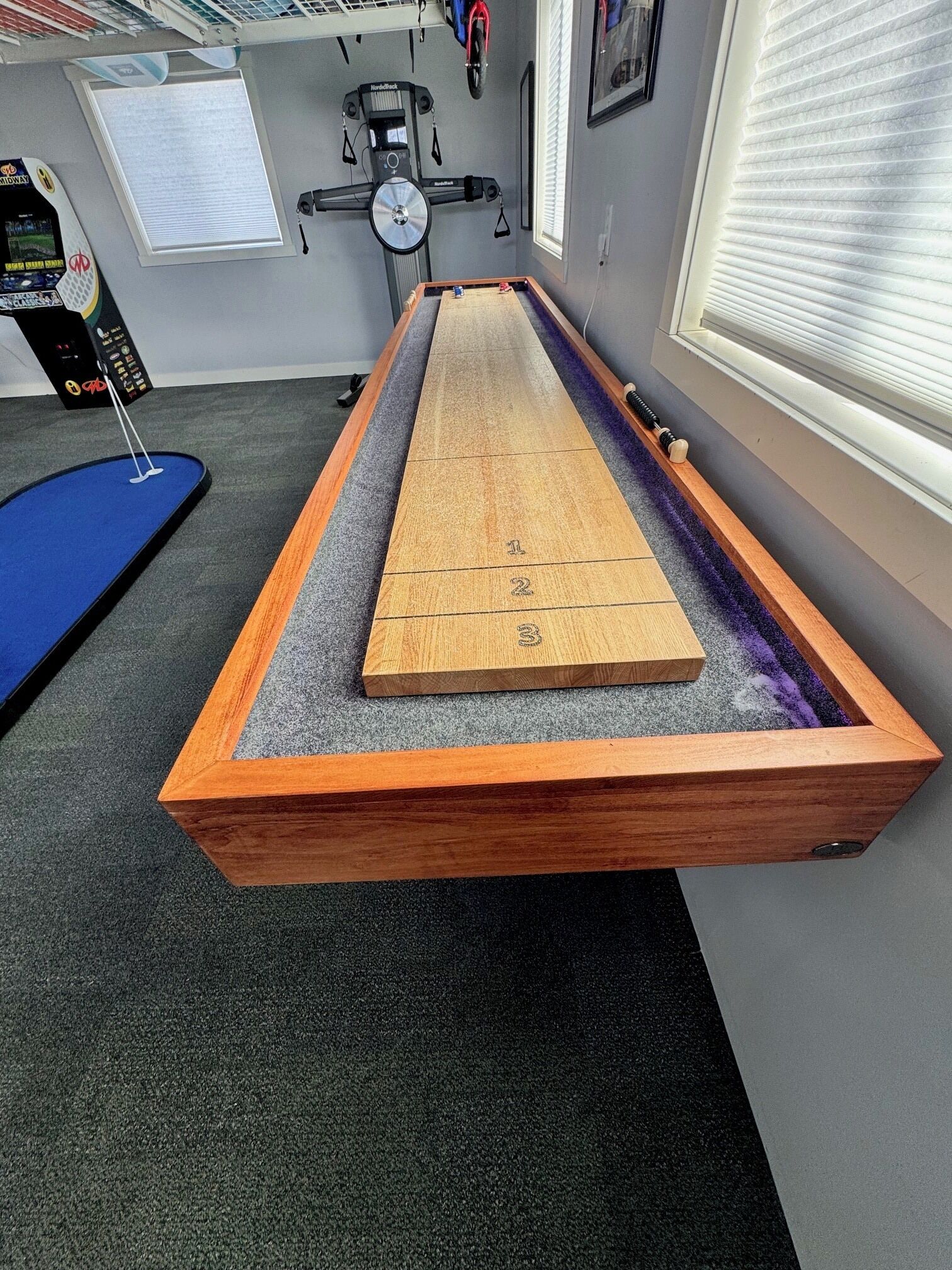 Ein Shuffleboard-Tisch mit nummerierter Spielfläche, ein Laufband und Trainingsgeräte im Hintergrund.