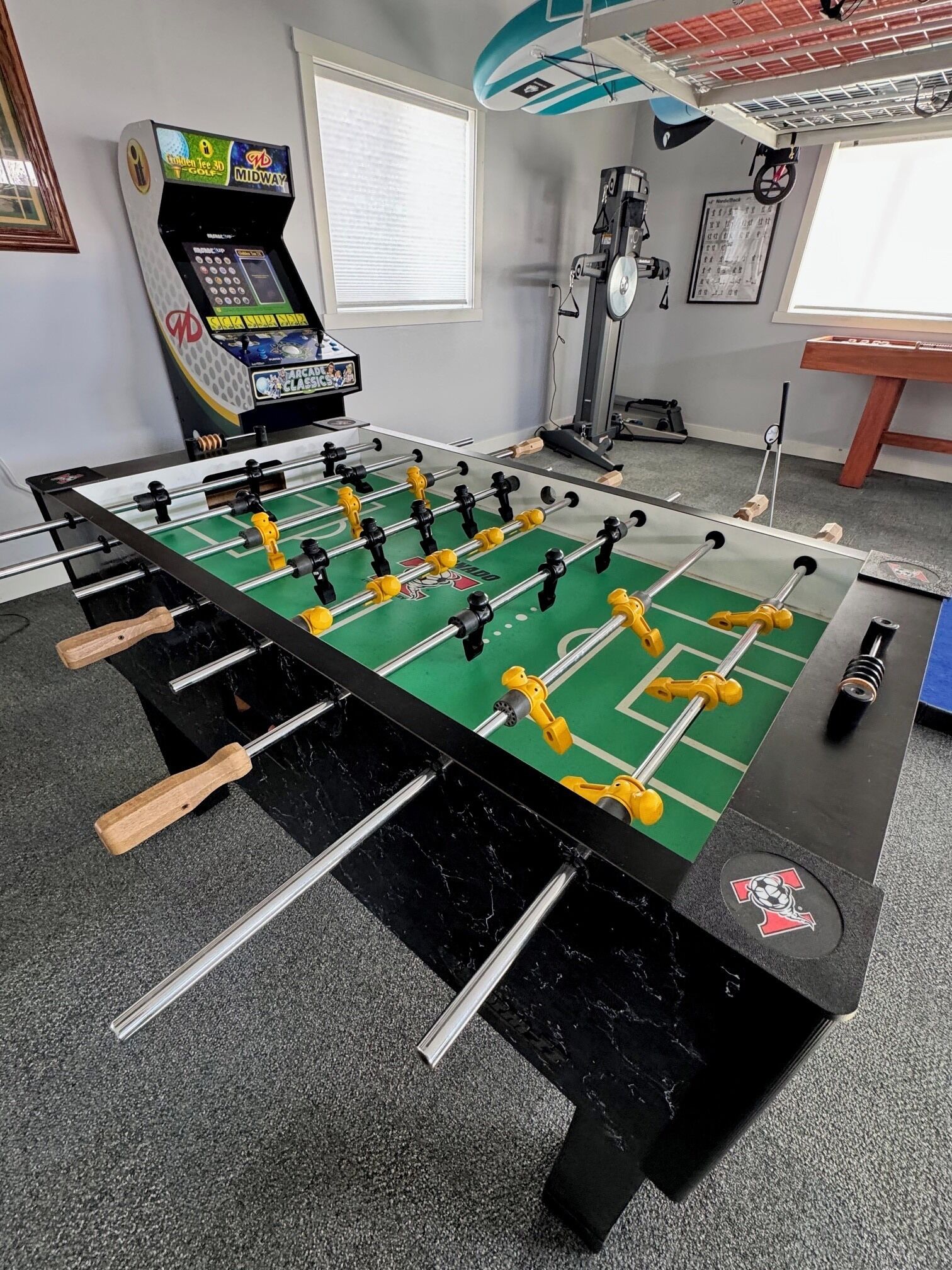 Una mesa de fútbol de mesa en una habitación con una ventana, una cinta de correr y una máquina de fútbol de mesa al fondo.